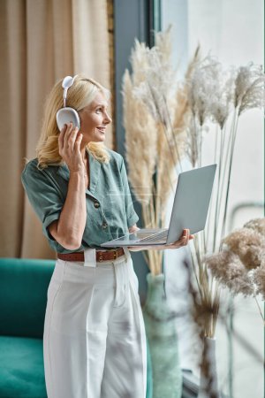 mujer positiva de mediana edad en auriculares inalámbricos escuchando música y utilizando el ordenador portátil en casa