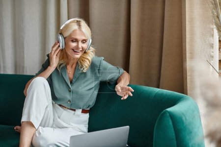 mujer de mediana edad en los auriculares inalámbricos sentado en el sofá y tener chat de vídeo en el ordenador portátil