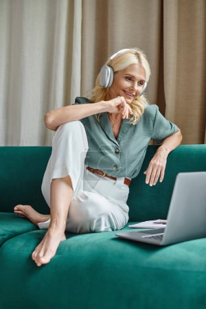glückliche Frau mittleren Alters in drahtlosen Kopfhörern, die auf dem Sofa sitzt und Laptop benutzt, von zu Hause aus arbeitet