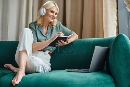 mujer de mediana edad positiva en auriculares inalámbricos sentados en el sofá cerca de la computadora portátil y portátil