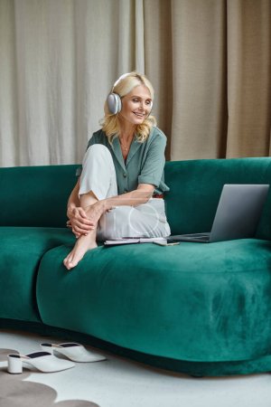 excited middle aged woman in wireless headphones sitting on sofa near laptop and listening webinar