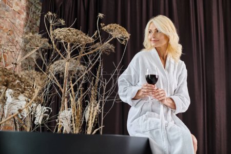 elegant blonde middle aged woman in white robe sitting near bathtub and holding glass of red wine