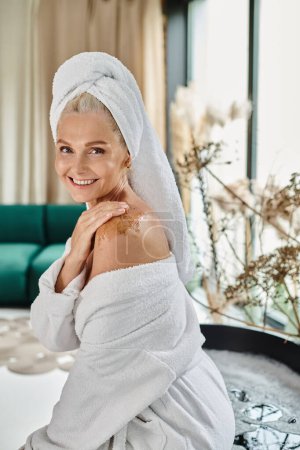 mujer alegre de mediana edad con toalla blanca en la cabeza y albornoz con exfoliación corporal en el baño