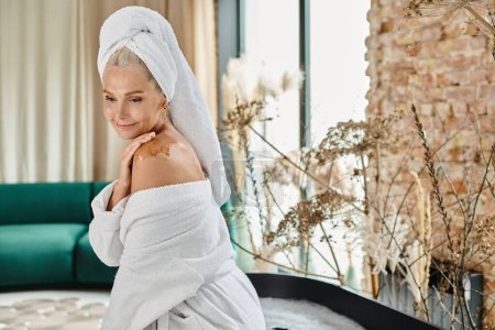 attractive middle aged woman with white towel on head and bathrobe using body scrub on shoulder