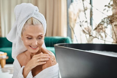 glückliche Frau mittleren Alters im weißen Bademantel und mit Handtuch auf dem Kopf, die Körperpeeling in der Nähe der Badewanne anwendet