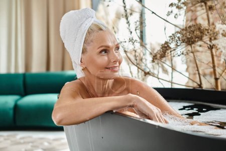 zufriedene Frau mittleren Alters mit weißem Handtuch auf dem Kopf badet in moderner Wohnung, Entspannung