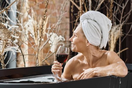 positive middle aged woman with towel on head holding glass of red wine while taking bath at home