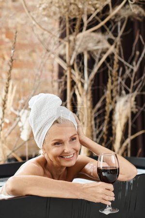 soñadora mujer de mediana edad con toalla en la cabeza sosteniendo un vaso de vino tinto mientras toma un baño