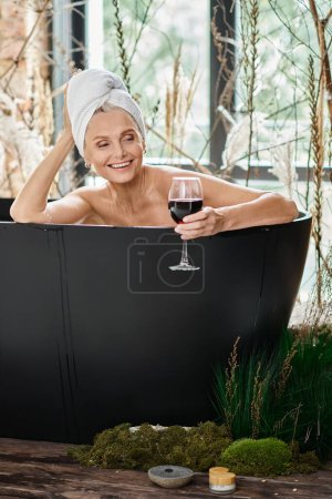 Foto de Sonriente mujer de mediana edad con toalla blanca en la cabeza sosteniendo un vaso de vino tinto y tomando un baño - Imagen libre de derechos