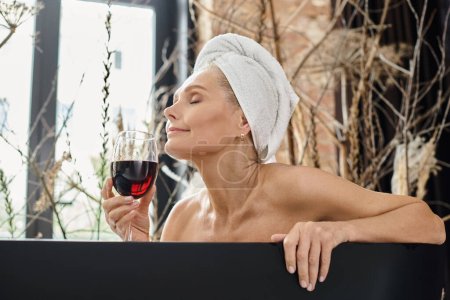 entspannte Frau mittleren Alters mit weißem Handtuch auf dem Kopf genießt den Geschmack von Rotwein beim Baden