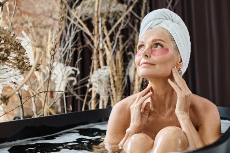 relaxed middle aged woman with white towel on head and under eye patches taking bath at home