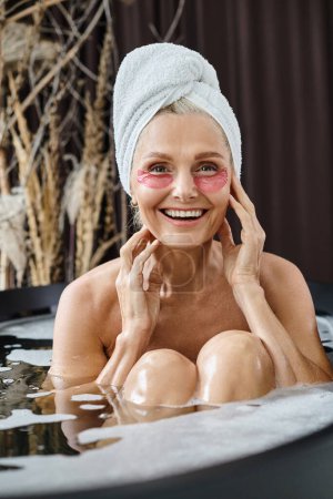 cheerful middle aged woman with white towel on head and under eye patches taking bath at home