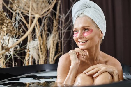 mujer alegre de mediana edad con toalla blanca en la cabeza y bajo parches para los ojos tomando un baño en casa