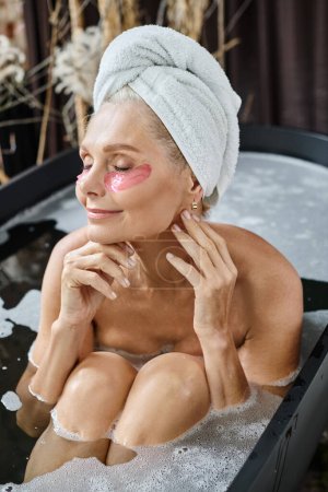 mujer de mediana edad complacida con toalla blanca en la cabeza y bajo los parches de los ojos tomando baño en casa