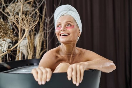 excited middle aged woman with white towel on head and under eye patches taking bath at home
