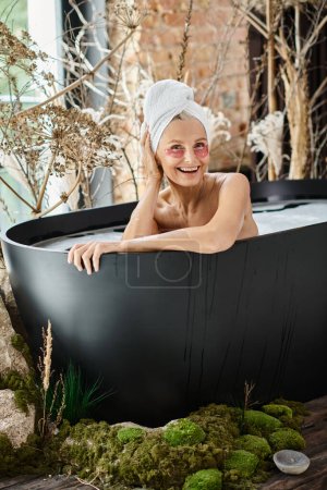 mujer radiante de mediana edad con toalla en la cabeza y bajo parches para los ojos hidratantes tomando un baño en casa