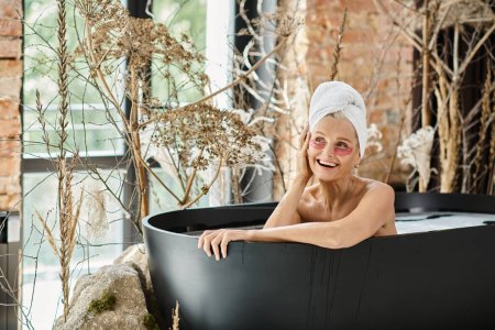 joyous middle aged woman with towel on head and eye patches taking bath in modern apartment