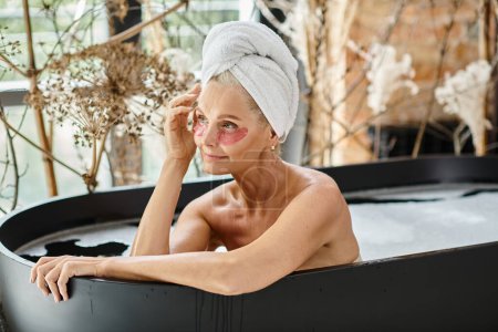femme d'âge moyen avec serviette sur la tête et des patchs hydratants pour les yeux prenant bain dans un appartement moderne