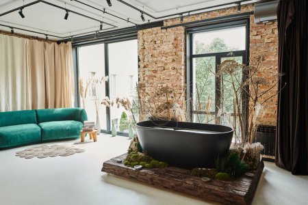 black bathtub inside of modern apartment with blue velour sofa, panoramic windows and plants