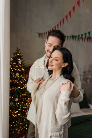 joyful man with closed eyes hugging happy wife in cozy home wear near blurred Christmas tree