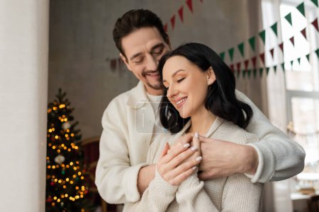 joyful man hugging wife in cozy home wear near blurred decorated Christmas tree, winter holidays