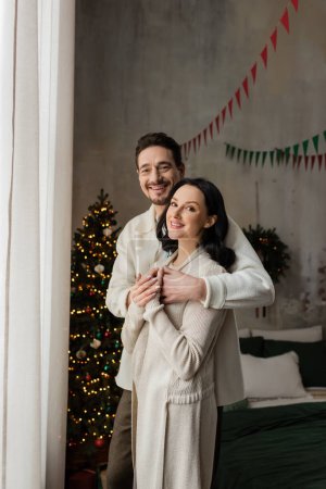 positive man hugging wife in cozy home wear near blurred decorated Christmas tree, winter holidays