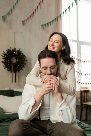 hombre cariñoso sentado en la cama y besar las manos de la esposa feliz cerca de la corona de Navidad en apartamento moderno