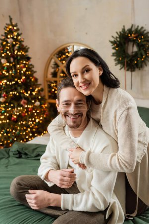 glückliche Frau umarmt bärtigen Mann neben verschwommenem Weihnachtsbaum in moderner Wohnung, Zufriedenheit