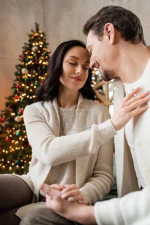 glückliche Frau mit geschlossenen Augen, die mit ihrem Mann in der Nähe des verschwommenen Weihnachtsbaums sitzt, Jahreszeit der Freude