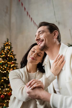 pleased woman with closed eyes sitting with husband near blurred Christmas tree, season of joy