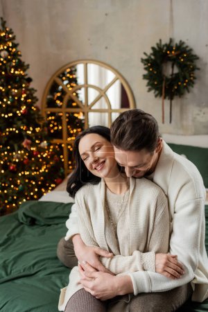 husband embracing joyful wife and sitting together on bed near blurred decorated Christmas tree
