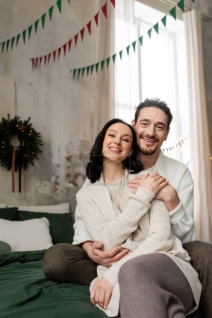 Mann umarmt fröhliche Frau und sitzt zusammen auf Bett neben verschwommenem Weihnachtskranz an Wand