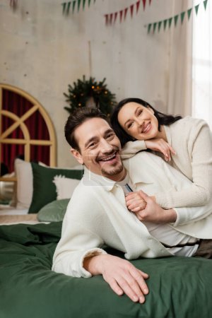 husband embracing cheerful wife and resting together on bed near blurred Christmas wreath on wall