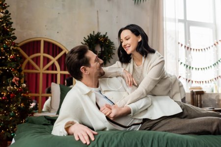 husband embracing cheerful wife and lying on bed near decorated Christmas tree and wreath on wall