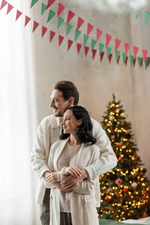 Glückliche Eheleute im Haus umarmen sich und stehen zusammen neben verschwommenem Weihnachtsbaum