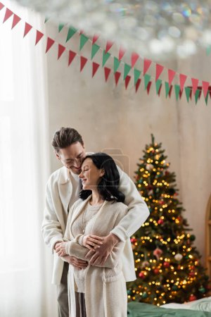 cheerful and married couple in home wear hugging and standing together near blurred Christmas tree