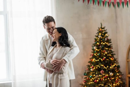 alegre y casado pareja en casa desgaste abrazo y de pie juntos cerca borrosa árbol de Navidad