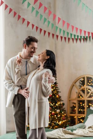 joyful husband and wife in home wear hugging and standing together near blurred Christmas tree