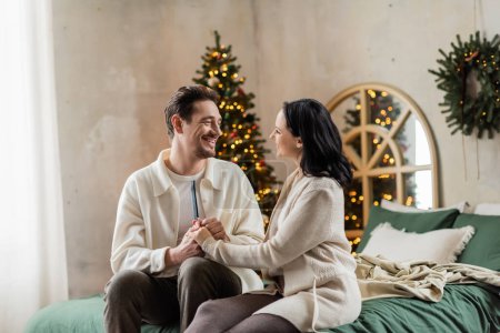 fröhliche Eheleute in Hauskleidung auf dem Bett sitzend und Händchen haltend in der Nähe des verschwommenen Weihnachtsbaums