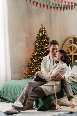 fröhliches Paar in Hauskleidung auf dem Bett sitzend und Händchen haltend neben verschwommenem Weihnachtsbaum zu Hause