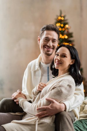 cozy living, joyful married couple hugging each other near blurred Christmas lights on backdrop