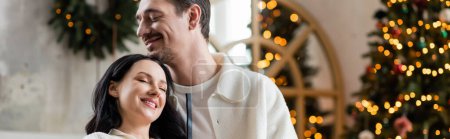happily married couple embracing near blurred lights of Christmas tree on backdrop, cozy banner