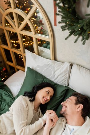 top view of joyful married couple spending cozy Christmas morning and looking at each other in bed