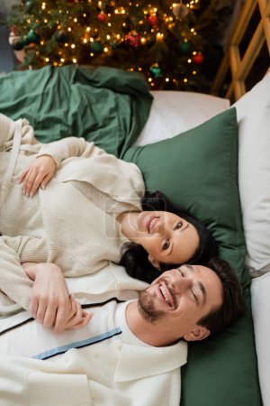 Ansicht von oben: freudiges Paar verbringt gemütlichen Morgen und liegt im Bett neben geschmücktem Weihnachtsbaum