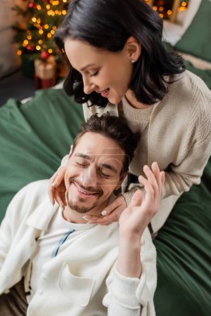 brunette woman hugging with husband and spending time together in bedroom near Christmas tree