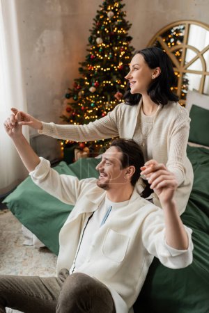 brunette woman holding hands of husband and spending time together in bedroom near Christmas tree