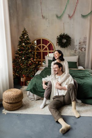 Winterurlaub, glückliche Frau umarmt Ehemann im geschmückten Schlafzimmer mit Weihnachtsbaum und Kranz