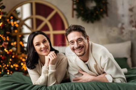 portrait of happy married couple looking at camera and lying together on bed near Christmas tree