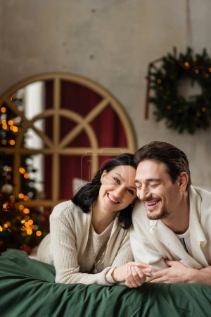 portrait of joyful married couple looking at camera and lying together on bed near Christmas tree