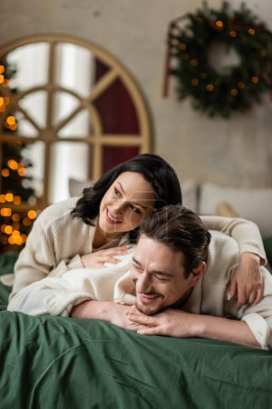 portrait of joyful married couple looking away and lying together on bed near Christmas tree
